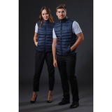 Two models wearing navy blue puffer vests, standing side by side against a dark background. The vests have a zip closure and a sleek design.