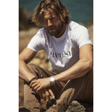 A white crew neck tee featuring a logo, worn by a man sitting outdoors with a rocky background.