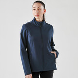 A woman wearing a navy softshell jacket with a high collar and side pockets, featuring a logo on the sleeve.