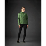 A woman stands wearing a green Women’s Kyoto Jacket paired with black leggings and shoes, with a dark background.