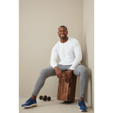 A man wearing a white long sleeve shirt sits on a wooden block, paired with grey pants and blue sneakers, with weights nearby.