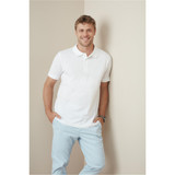 A man wearing a white premium cotton polo shirt, smiling while leaning against a wall, paired with light blue trousers.