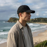 A black linen cap is worn by a person on a beach, with ocean waves and cliffs in the background.