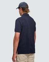A man wearing a navy ultrafine cotton pique polo shirt, viewed from the back, paired with light-coloured trousers.