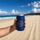 A drink bottle holder in navy blue, featuring a logo, held against a beach backdrop with ocean waves.