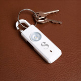 A white safety alarm keychain with a circular light, attached to a set of silver keys on a brown leather surface.