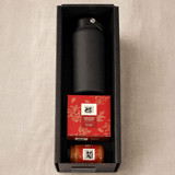 A black drink bottle, a red box of Christmas pudding, and a jar of golden sauce in a black box.