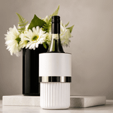 A white wine bottle cooler with a silver band, placed on a marble surface beside a black vase with white flowers.