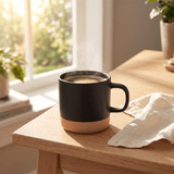 A black and terracotta ceramic coffee mug with steam rising, placed on a wooden table near a window.