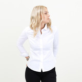 A white women's shirt with long sleeves and a button-up front, worn with black trousers. The model stands facing sideways.