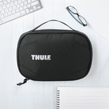 A black toiletry bag with a handle and a logo on the front, placed on a light wooden surface with office supplies.