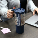 A drink bottle featuring a blue body and decorative patterned sleeve, with a handle and straw, resting on a table.