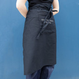 A black long waist apron with a side pocket, featuring a logo on the front.