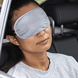 A grey eye mask rests over a woman's eyes as she relaxes in a car seat, featuring a logo on the mask.