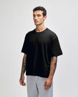 A black unisex boxy t-shirt made of heavyweight cotton, displayed on a model with tattoos, paired with light grey pants.