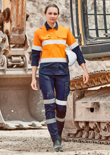 A woman wearing orange and navy cargo pants with reflective stripes, standing near heavy machinery. A hard hat is on her head.
