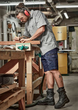 A man in a grey shirt and navy shorts, wearing safety gear, works at a woodworking bench in a workshop.