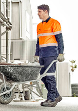 A man in an orange and navy utility pant pushes a wheelbarrow at a construction site. The pants have reflective detailing and a logo.