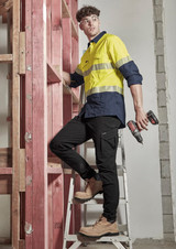 A man in a yellow and navy long sleeve shirt stands on a ladder, holding power tools, next to timber framing.