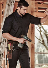 A man in a black rugged long sleeve shirt is holding a power drill, standing inside a construction site.