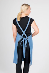 A blue cotton/denim apron with cross-back straps, featuring a tie at the waist and an eyelet detail.