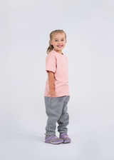 A child wearing a pink t-shirt and grey pants, smiling with a neutral background. A logo is visible on the shirt.