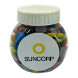 A clear plastic jar filled with colourful choc beans, featuring a white lid and a label with a logo.
