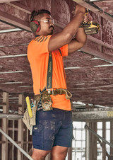 A man wearing an orange shirt and navy blue cargo shorts works on a construction site, using a power drill.