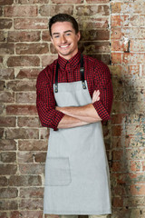 A grey bib apron with adjustable straps, worn over a red and black checkered shirt, against a brick wall background.