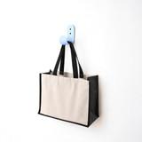 A functional tote bag in beige and black hangs on a blue hook against a white wall. The bag has handles and a logo.