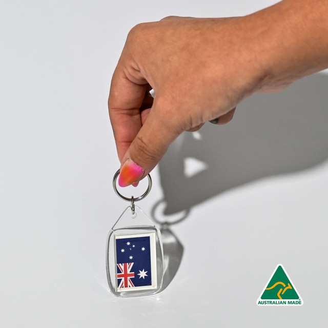 A hand holds a clear tear drop acrylic keychain featuring the Australian flag. The keychain has a silver ring and a logo.