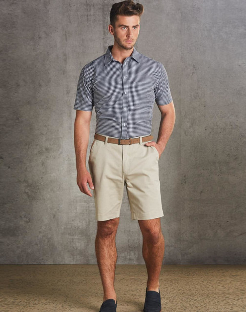 Men's chino shorts in beige, paired with a checked short-sleeve shirt and a brown belt. The model stands against a textured grey backdrop.