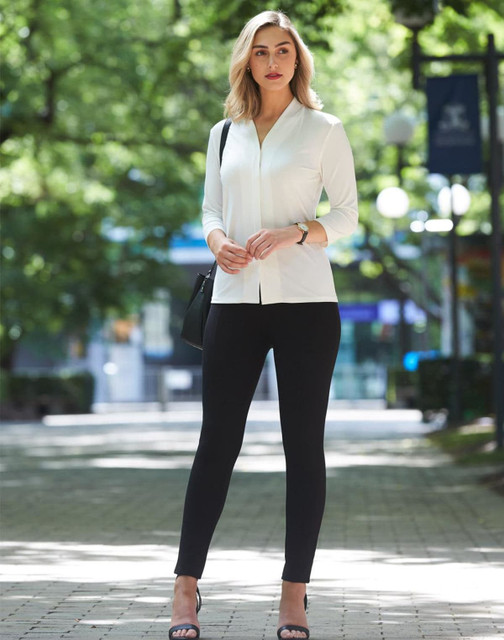 A model stands outdoors wearing black treggings and a white top, complemented by heels and a handbag.