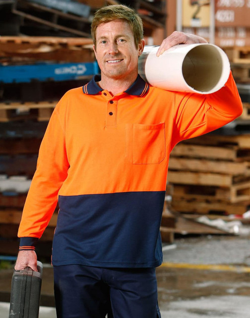 A man poses in an orange and navy long-sleeve safety polo shirt, holding a pipe and toolbox in a work environment.