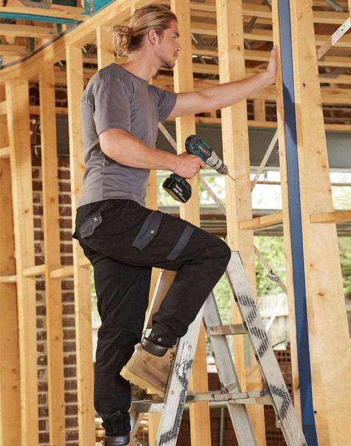 A pair of black work pants on a model using a power drill while standing on a ladder amidst wooden framing.