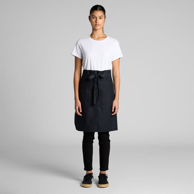 A half apron in black, featuring a front pocket and a tied waistband, displayed on a model wearing casual attire.