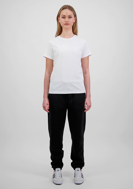 A woman in a white t-shirt and black track pants stands against a plain background. The pants have a branding logo.