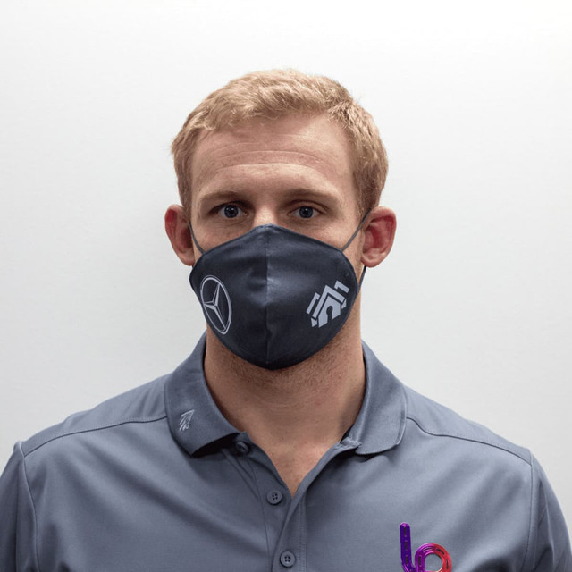 A grey face mask featuring a logo, worn by a person with short light hair and a grey shirt.