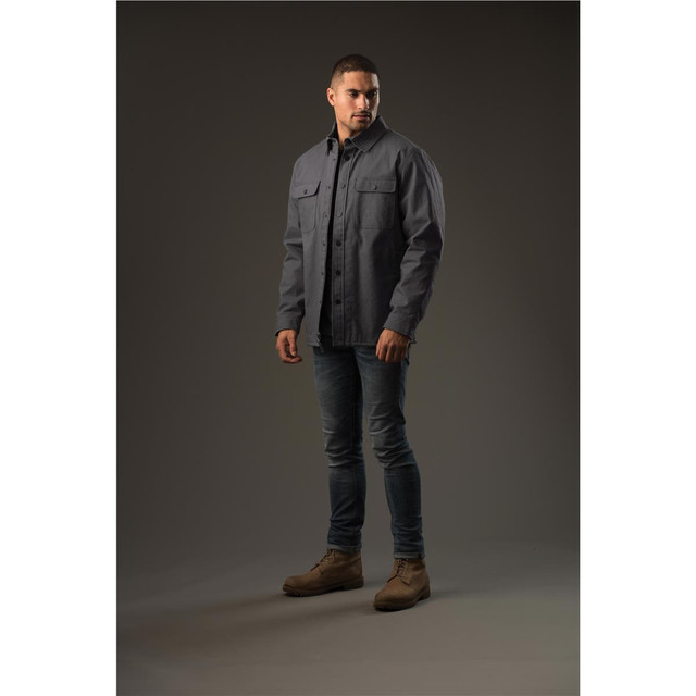 A man stands wearing a grey Men's Tradesmith Jacket, styled with fitted jeans and tan boots. The background is neutral.