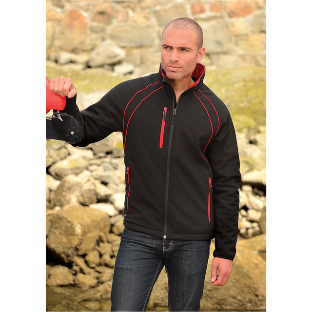 A man wearing a black softshell jacket with red accents, standing outdoors by rocky terrain. The jacket has a logo.