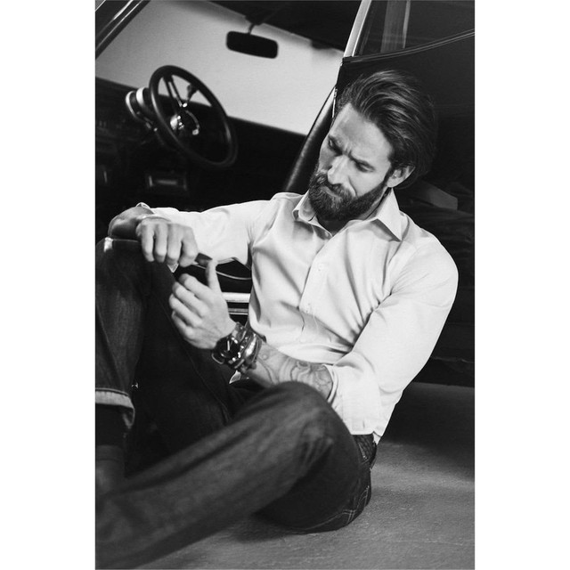 A man wearing a green button-up shirt sits on the ground, looking at his wrist, with a vintage car in the background.