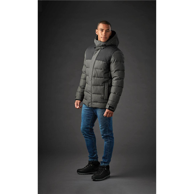 A men's parka in dark grey with a hood, featuring a quilted design and worn over denim jeans and boots.
