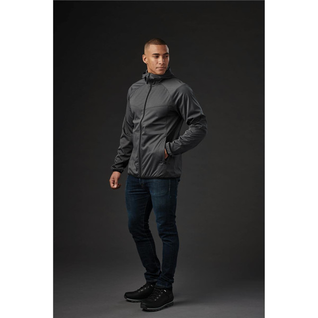 A model wearing a grey men's softshell jacket, paired with dark jeans and black shoes, standing against a dark background.