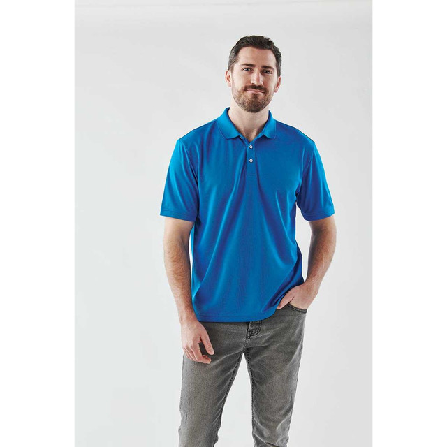 A man wearing a bright blue polo shirt with a collar and a logo on the chest, paired with grey trousers.