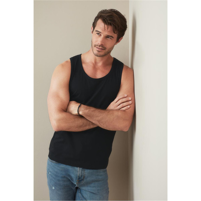 A men's classic tank top in black, displayed by a model leaning against a wall, wearing jeans.