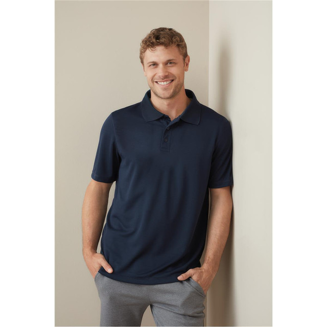 A man wearing a navy blue polo shirt stands against a neutral wall, smiling and casually posing.