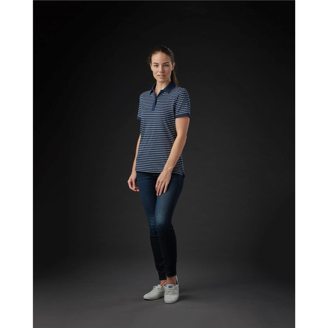 A woman wearing a navy and white striped polo shirt, paired with blue jeans and white shoes, standing against a dark background.