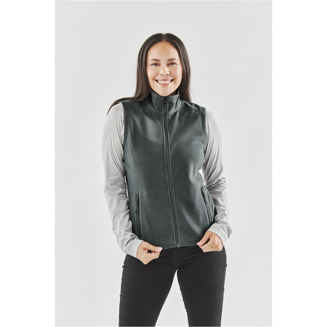A woman stands wearing a dark green fleece vest over a light grey long-sleeve top, smiling at the camera.