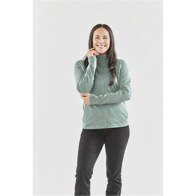A woman wearing a light green zip-up performance jacket, standing with a smile, paired with black pants.