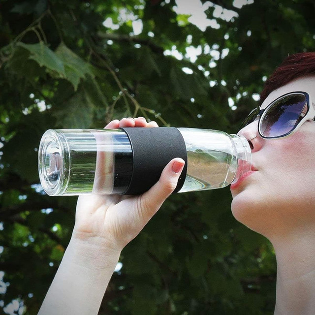 A drink bottle with a clear body and a black silicone grip, held by a person sipping water outdoors.